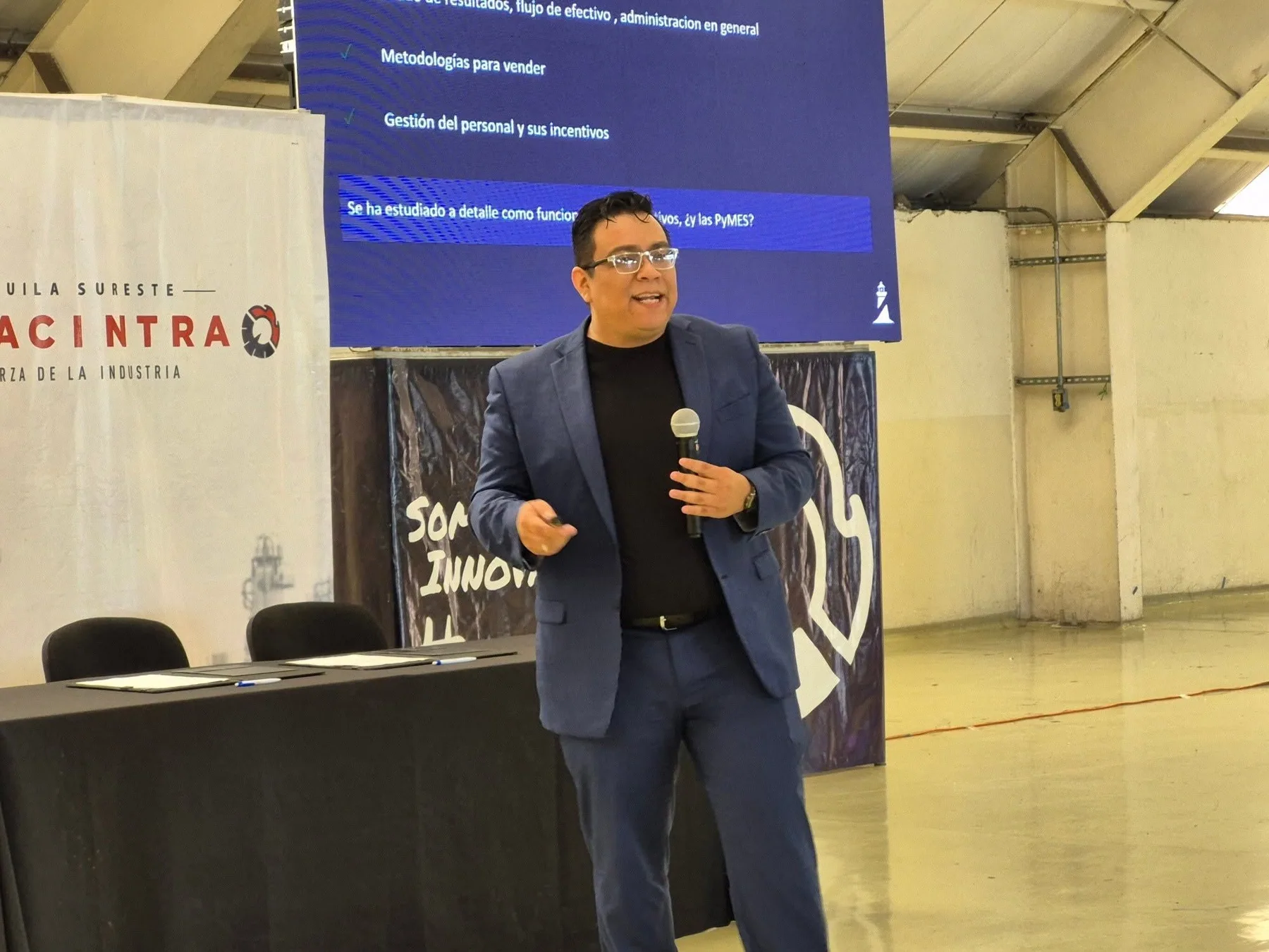 Alan con saco azul participa con un micrófono frente a una pantalla durante un evento.