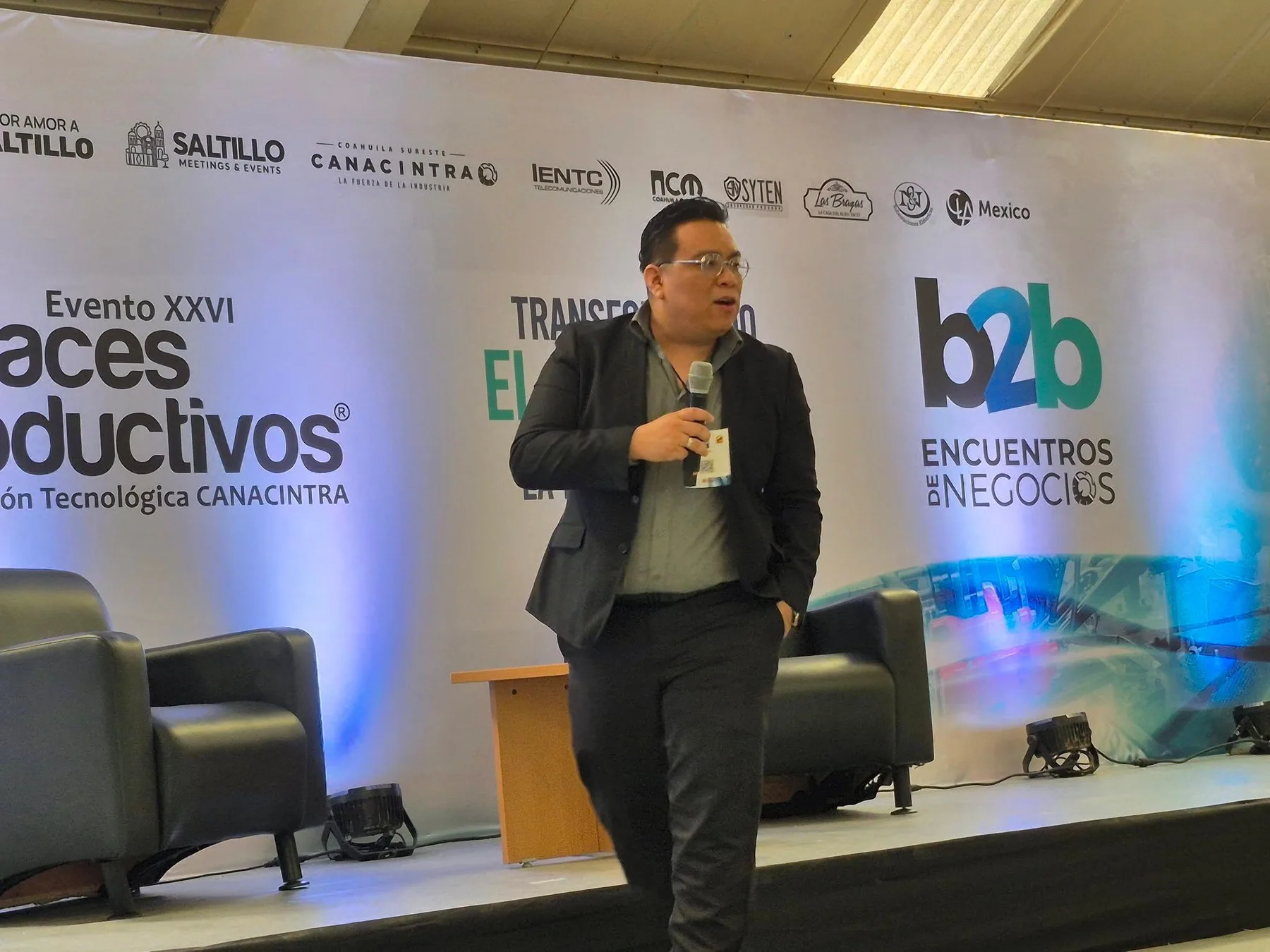 Alan habla con un micrófono en un escenario frente a una pantalla de evento.