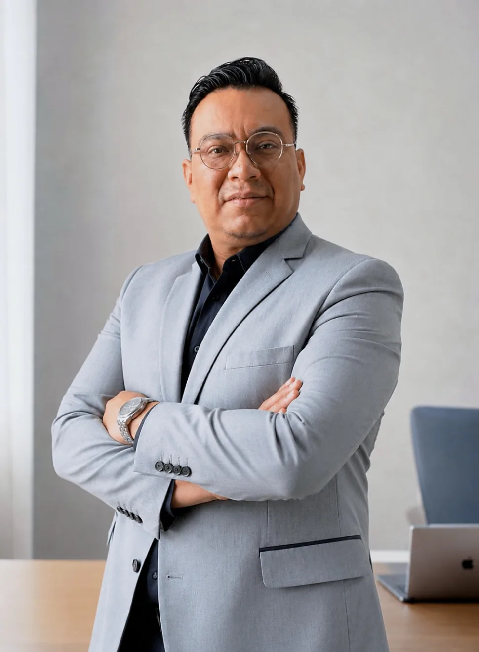 Alan Guillén con saco gris y camisa negra, brazos cruzados; una computadora se ve al fondo.